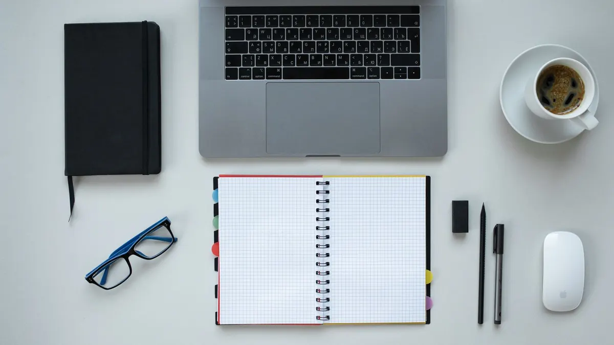 Opgeruimde thuiswerkplek met laptop, notitieboek, bril en kop koffie op een wit bureau